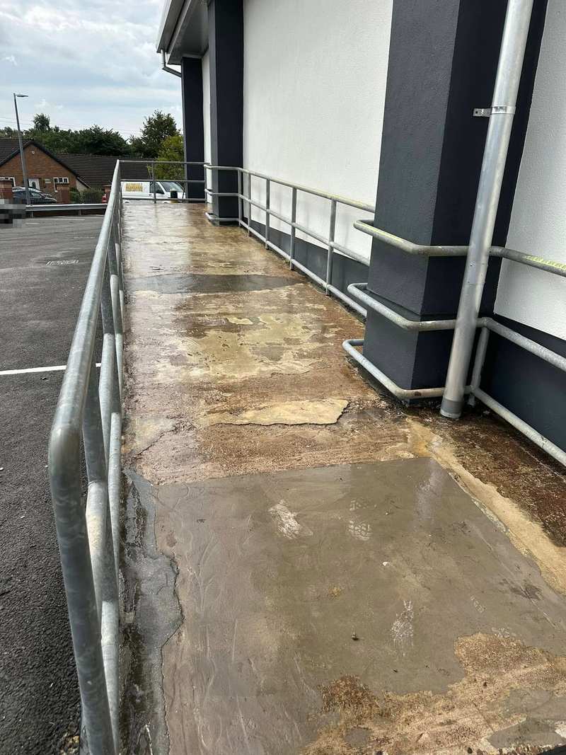 Worn, stained concrete access ramp with galvanised handrails beside a supermarket wall