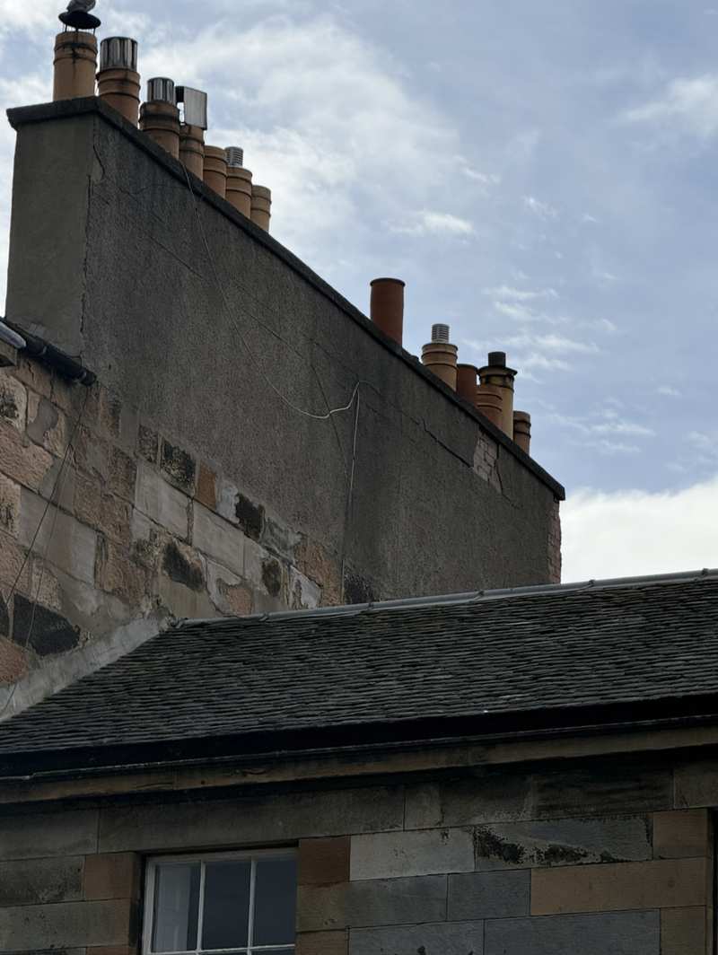 Long rendered chimney stack with cracked, weathered render and a row of clay chimney pots, above a slate roof and stone wall