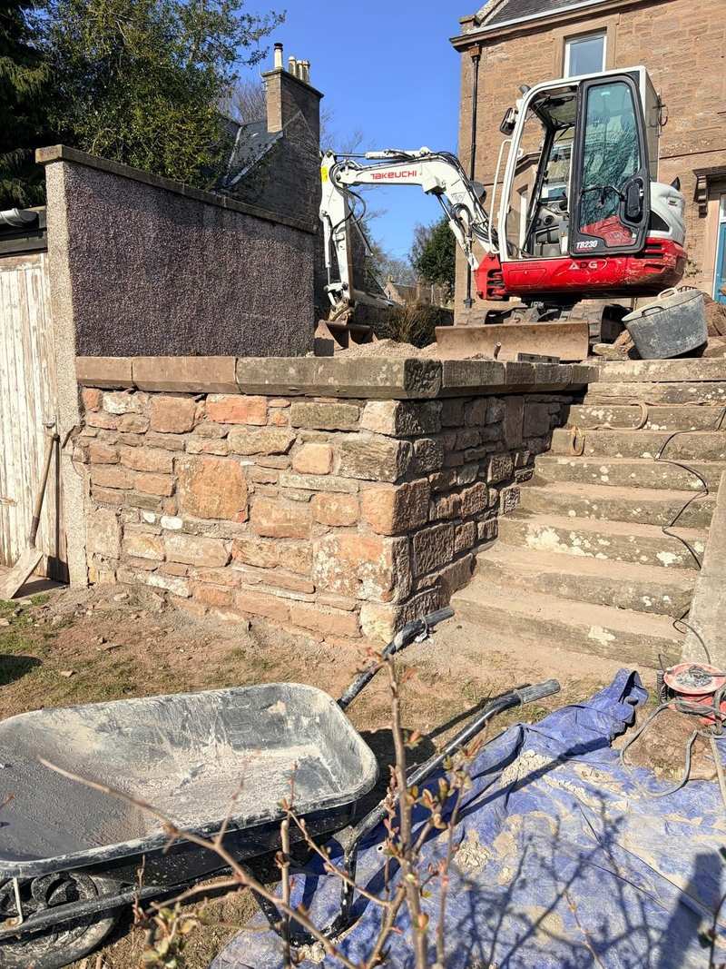 Rebuilt red sandstone boundary wall with new stone coping beside a flight of stone steps, with a mini excavator behind